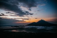 Serene view of Mount Batur silhouetted against a colorful sunset sky, surrounded by clouds and reflecting water.